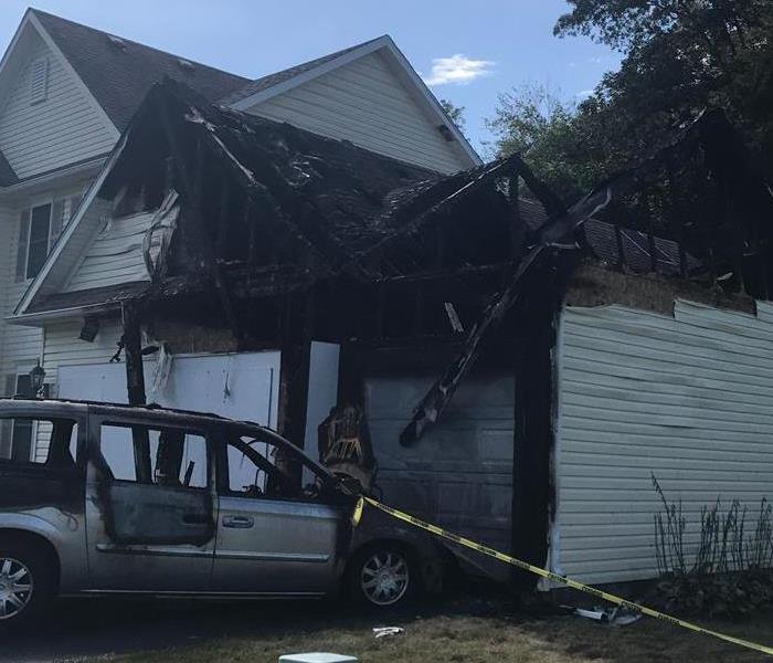 A home with severe fire damage to the garage with a hole in the roof and a burned car.